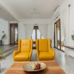 4 BHK Villas Elements Nachinola Goa living area with yellow accent chairs wooden coffee table patterned flooring white curtains and open layout design